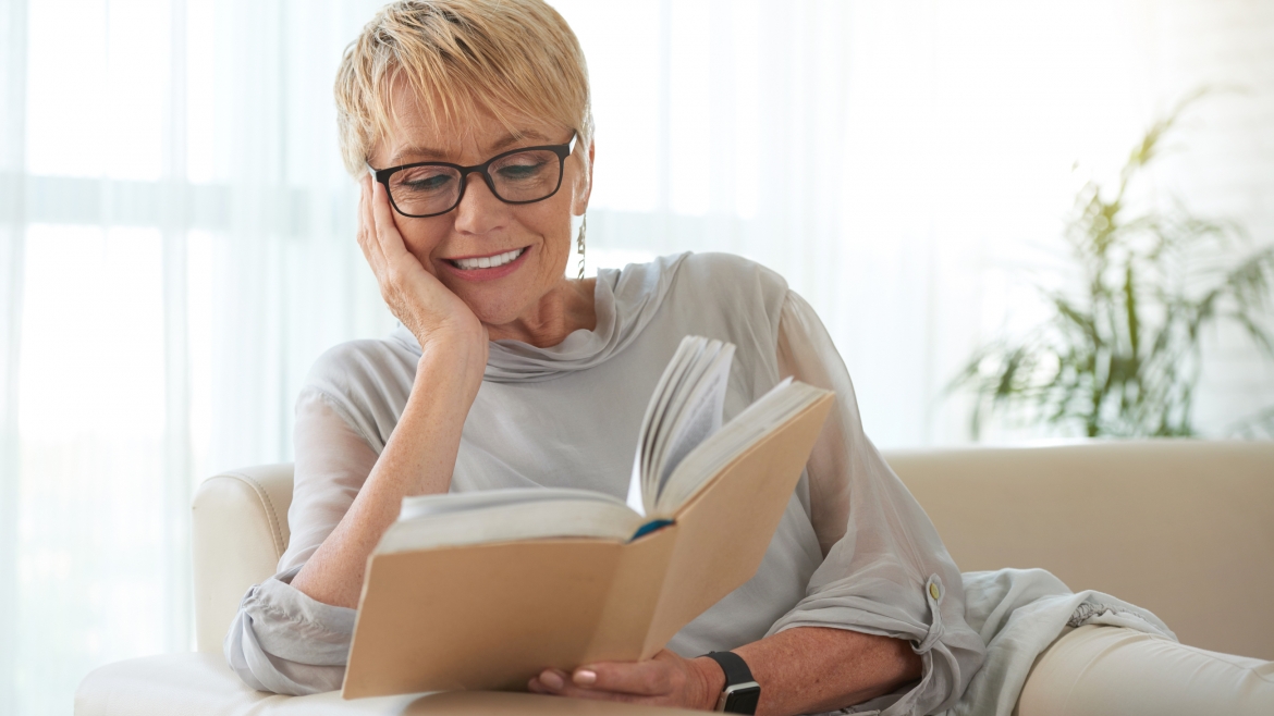 Senior blond woman in glasses resting on sofa and reading a book