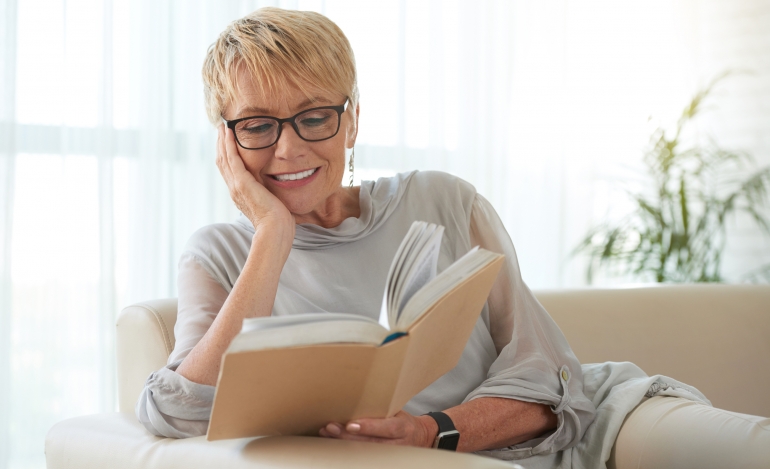 Senior blond woman in glasses resting on sofa and reading a book